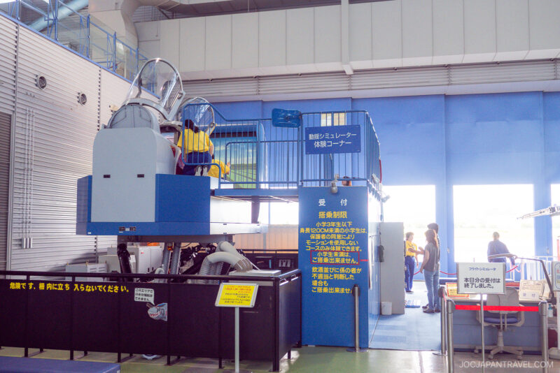 A flight simulator setup indoors at Airpark Hamamatsu features a cockpit replica elevated on a blue platform, with people in yellow shirts interacting around it. Japanese signs and informational panels are visible in the bright, spacious room.