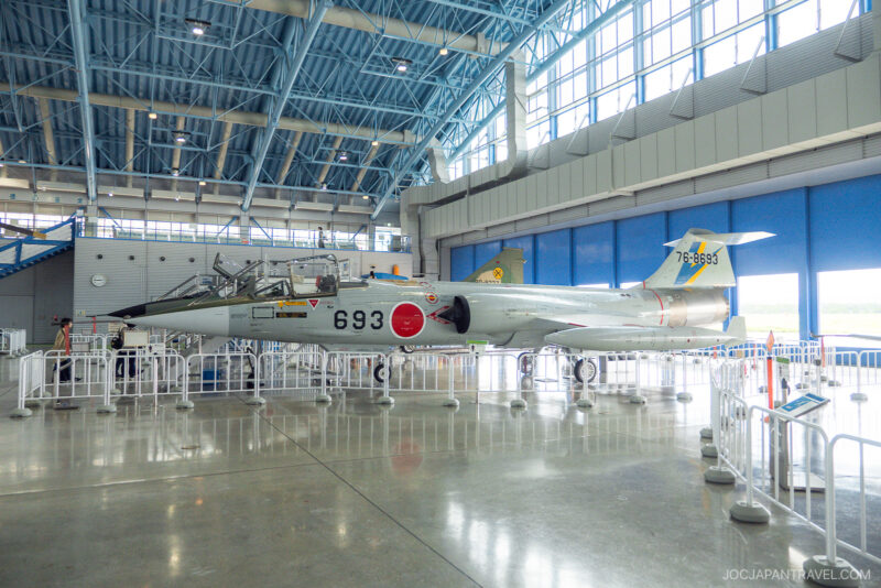 A silver fighter jet with the number 693 and Japanese markings is displayed inside the spacious, modern Airpark Hamamatsu museum hangar with blue steel beams and large windows, surrounded by white railings.