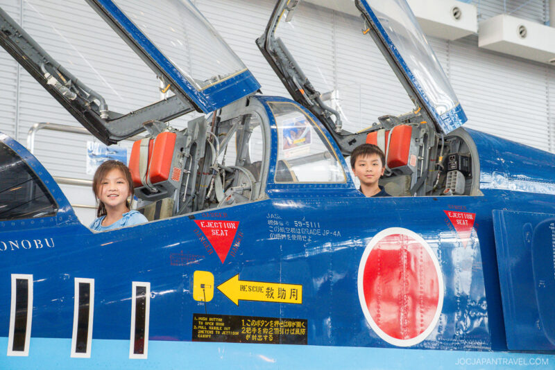 Two children sit inside the open cockpits of a blue military jet at Airpark Hamamatsu, smiling at the camera. The jet features Japanese writing, warning labels, and a red circle insignia on its side.