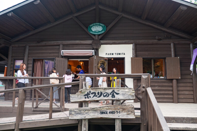 Visitors gather outside and enter Farm Tomita, a rustic wooden building with a Japanese sign on the deck, surrounded by lush greenery on a cloudy day.