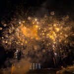 Fireworks in Izunokuni (伊豆の国市) – Summer Festival Magic Bright golden fireworks burst in the night sky above silhouetted buildings at the Izunokuni Fireworks Festival, with smoke drifting across the scene. The lights illuminate the dark, festive atmosphere below.