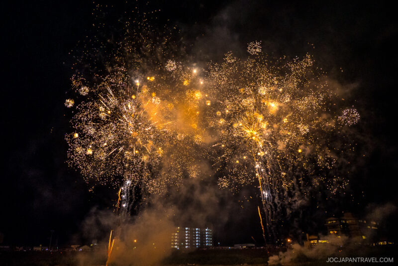 Bright golden fireworks burst in the night sky above silhouetted buildings at the Izunokuni Fireworks Festival, with smoke drifting across the scene. The lights illuminate the dark, festive atmosphere below.
