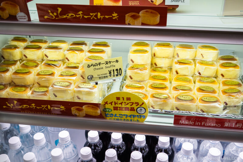 Cheese soufflé cakes from Furano Cheese Factory are displayed in clear plastic containers with price tags in Japanese and English. Bottled water is visible on the shelf below.