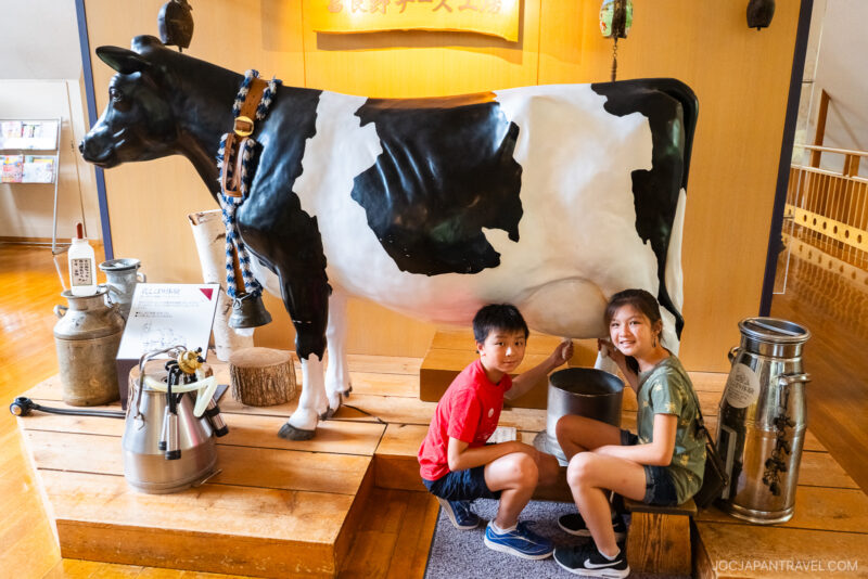 Two smiling children kneel beside a life-sized black and white cow statue indoors at the Furano Cheese Factory, pretending to milk it. Various milk cans and buckets are displayed nearby on wooden platforms.