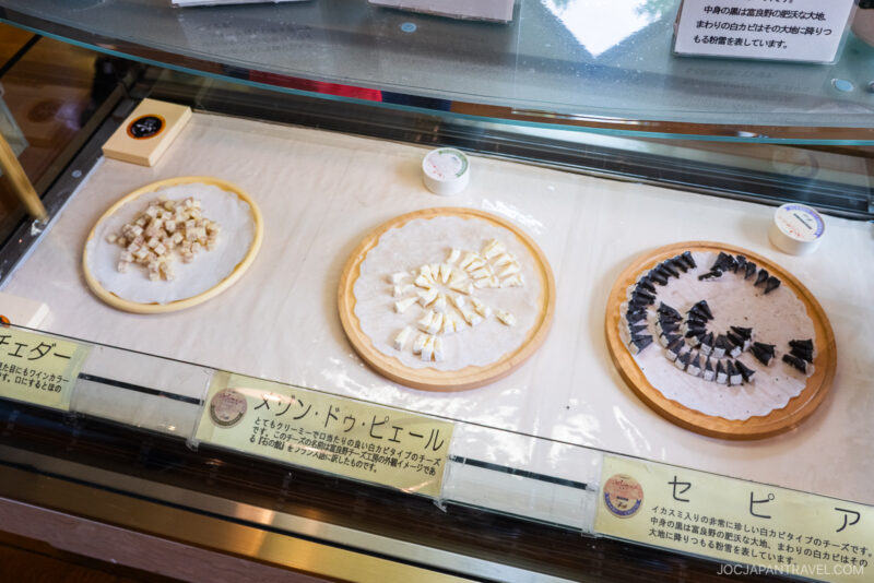 Three pizza crusts are displayed on a counter, each topped with different cut ingredients for sampling. The setup, featuring Japanese and English labels, showcases offerings from the Furano Cheese Factory in an inviting food display.