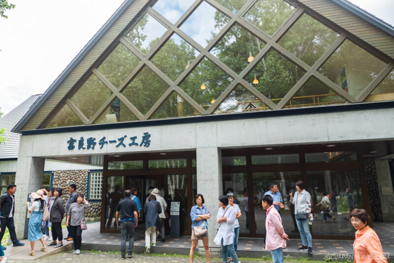 A group of people stands outside a modern building with large windows and a triangular roof. The sign above the entrance reads Nara Cheese Factory in Japanese, reminiscent of the iconic Furano Cheese Factory. Trees are reflected in the glass.