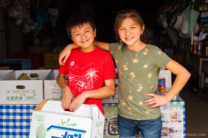 Two smiling children stand close together in front of a market stall with boxes of produce, including Furano Melon. The boy wears a red t-shirt and the girl, in a green shirt with a palm tree pattern, has her arm around his shoulders.