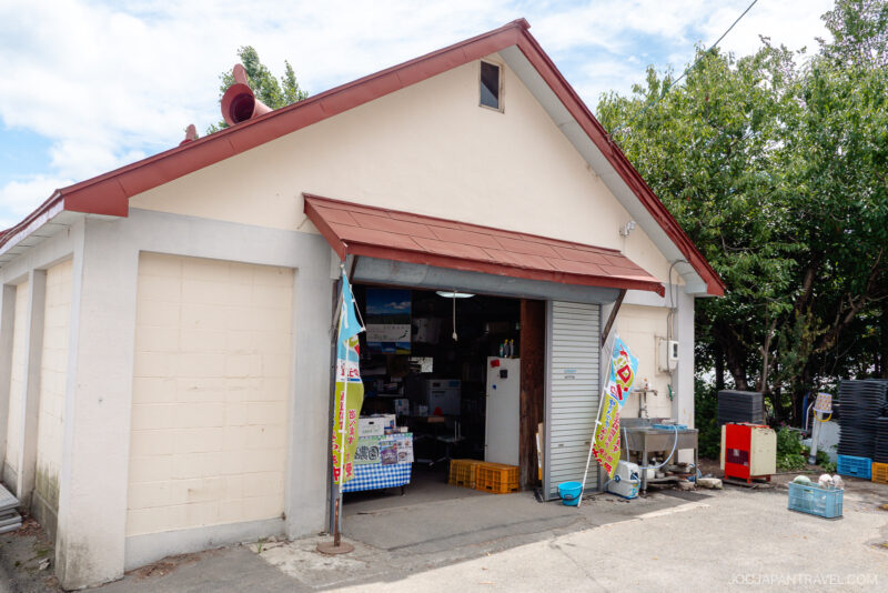 A small, simple shop with a red roof and an open garage-style door displays banners and vending machines outside. Inside, various goods and a refrigerator are visible—perhaps even some famous Furano Melon. Trees and blue sky form the backdrop.