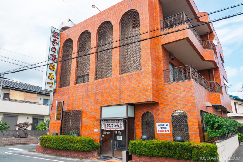 A three-story building with orange brick exterior, arched windows, and metal railings; Japanese signs and a Hamamatsu Gyoza banner are displayed on the facade. The building sits on a quiet street with greenery around it.