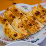 Hamamatsu Gyoza (浜松餃子) – Japan’s Cabbage-Filled Dumpling Specialty A close-up of eight golden-brown Hamamatsu Gyoza dumplings arranged in a row on a decorative white plate with red patterns.