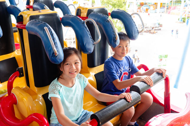 Two children sit side by side in seats on a Hamanako Pal Pal amusement park ride, smiling at the camera. The ride features bright yellow and red colors with blue safety harnesses above their heads.