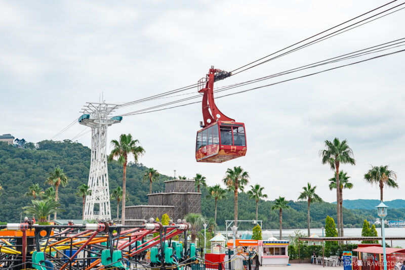 A red cable car travels along suspended cables above Hamanako Pal Pal amusement park, with palm trees, colorful rides, and a distant view of green, forested hills and water.