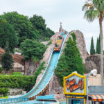 A water ride boat descends a steep blue track on a rocky hill surrounded by greenery at Hamanako Pal Pal amusement park. The Wild River sign is visible in the foreground near palm trees and other park structures.