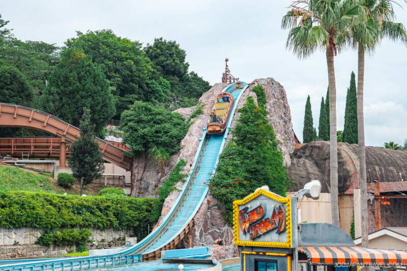 A water ride boat descends a steep blue track on a rocky hill surrounded by greenery at Hamanako Pal Pal amusement park. The Wild River sign is visible in the foreground near palm trees and other park structures.