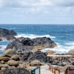 Hirauchi Kaichu Natural Onsen (平内海中温泉) – A Tidal Hot Spring Experience in Yakushima Rocky coastline with waves crashing against dark rocks under a partly cloudy sky. In the foreground, a wooden walkway and sign lead toward Hirauchi Kaichu Natural Onsen, with the ocean stretching out in the background.