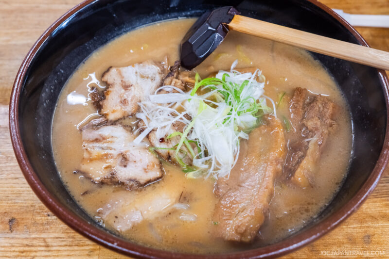 A bowl of Sapporo Ramen with slices of roast pork, green onions, and noodles in a rich, creamy broth, served with a wooden spoon on a wooden table.