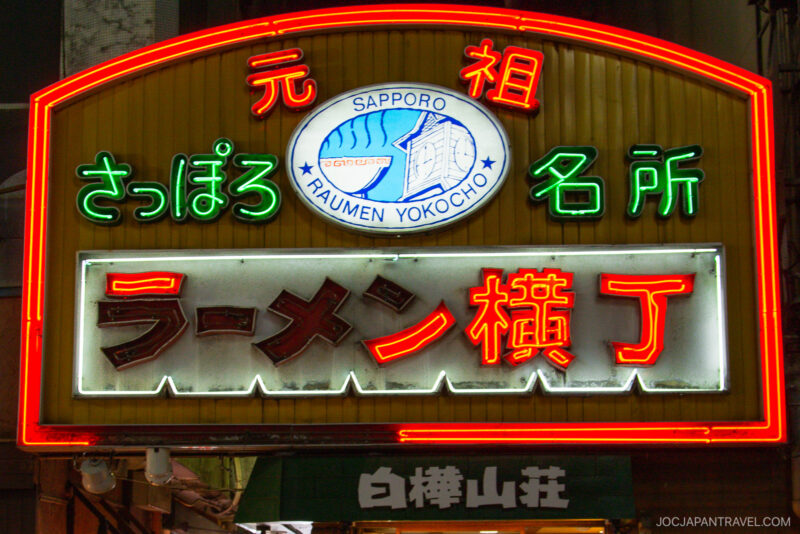 A brightly lit neon sign at night for “Sapporo Ramen Yokocho” in Japanese and English marks this famous Sapporo Ramen alley, glowing with green, red, and white lights.
