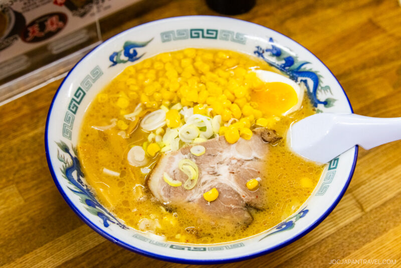 A bowl of Sapporo Ramen with corn, sliced green onions, a piece of pork, and a soft-boiled egg in a rich broth, served in a decorative blue and white bowl with a white spoon.