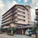 Hotel Tachibana Shikitei, a multi-story traditional Japanese ryokan with wooden balconies and a tiled roof, stands beside a quiet street with trees and a white van parked at the entrance under a partly cloudy sky.