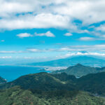 A scenic view of lush green hills at Izunokuni Panorama Park leads to a bright blue sea under a partly cloudy sky, with a distant mountainous coastline visible on the horizon.