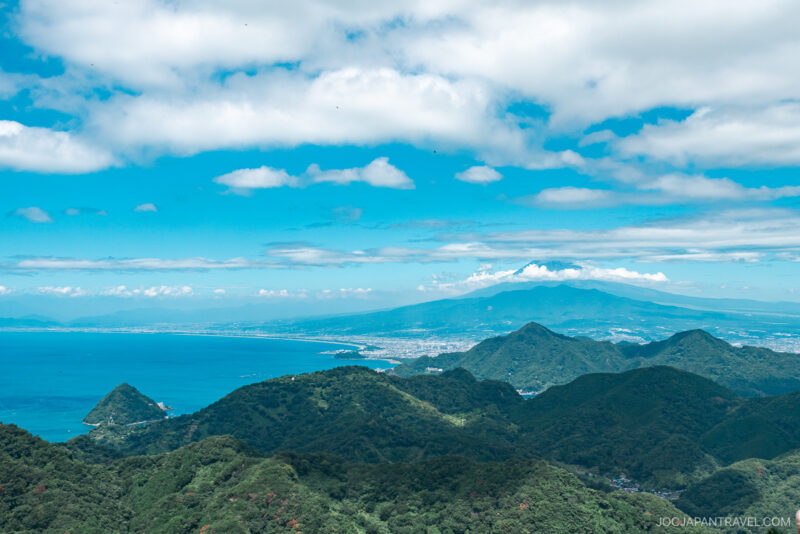 A scenic view of lush green hills at Izunokuni Panorama Park leads to a bright blue sea under a partly cloudy sky, with a distant mountainous coastline visible on the horizon.