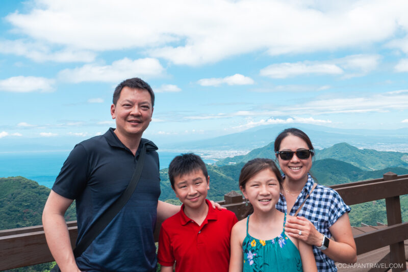 A family of four smiles on a wooden observation deck at Izunokuni Panorama Park, with mountains, green hills, and a blue sky with clouds in the background. The parents stand behind their two children, enjoying the scenic view.