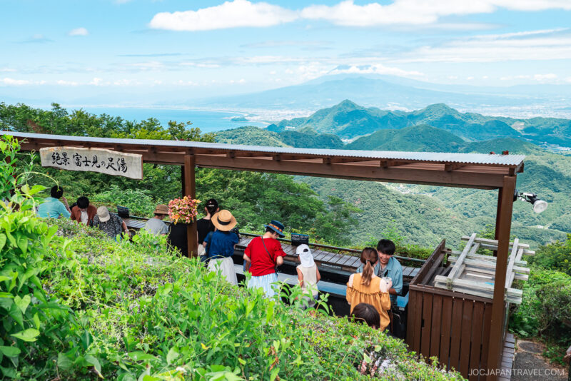 A group of people relax at an outdoor footbath in Izunokuni Panorama Park on a mountain, surrounded by lush greenery, enjoying a panoramic view of hills, a distant city, and the sea under a blue sky.