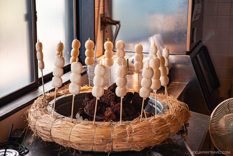 Skewers of Japanese dango rice dumplings are arranged in a circle around a charcoal grill, set inside a straw-wrapped base at Izunokuni Panorama Park, with light streaming through a nearby window.