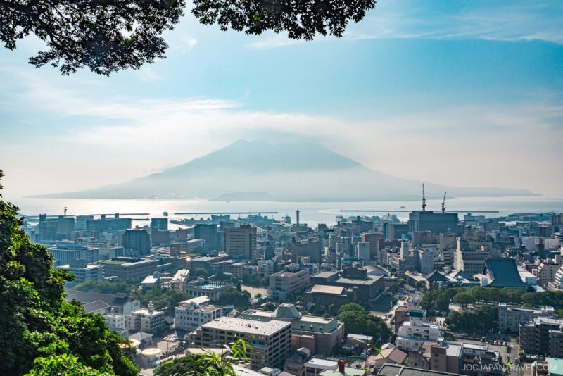 View of a coastal city with a prominent volcano, Sakurajima, rising in the background across the water under a hazy sky, framed by tree branches—Kagoshima shopping awaits just beyond this picturesque scene.