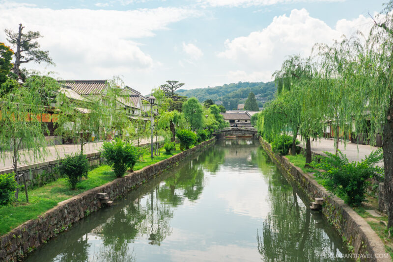 A peaceful canal lined with green trees and traditional Japanese buildings on both sides—discover this tranquil scene in our Kurashiki travel guide, featuring a small stone bridge beneath a partly cloudy sky.