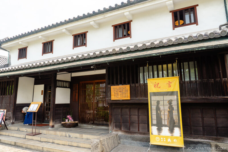 Traditional Japanese building with white plaster walls, dark wooden beams, and tile roof. A Kurashiki Travel guide is displayed by the entrance, surrounded by potted plants, creating a calm and inviting scene.