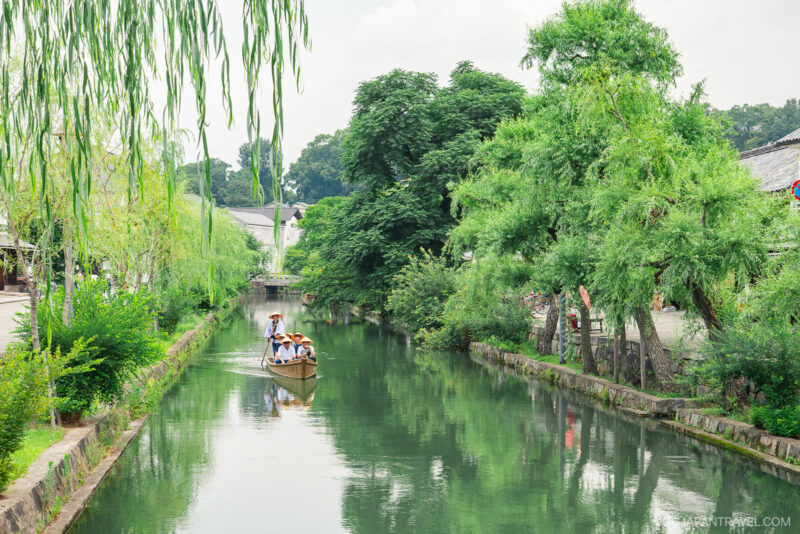 A small wooden boat with several people is being rowed along a calm, narrow canal lined with lush green trees and willow branches on a bright day in Kurashiki. Traditional buildings complete this picturesque scene from any Kurashiki travel guide.