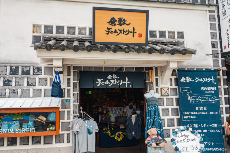 A Japanese denim shop with a traditional-style exterior, clothing displayed outside, and a large wooden sign with Japanese text above the entrance—an authentic spot featured in many Kurashiki travel guides. Maps and shirts hang on the wall beside the door.