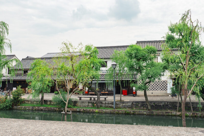 Traditional Japanese buildings with tiled roofs stand behind a canal, bordered by green trees and a stone pathway under a cloudy sky. Benches and a street lamp near the water complete this serene Kurashiki Travel guide scene.