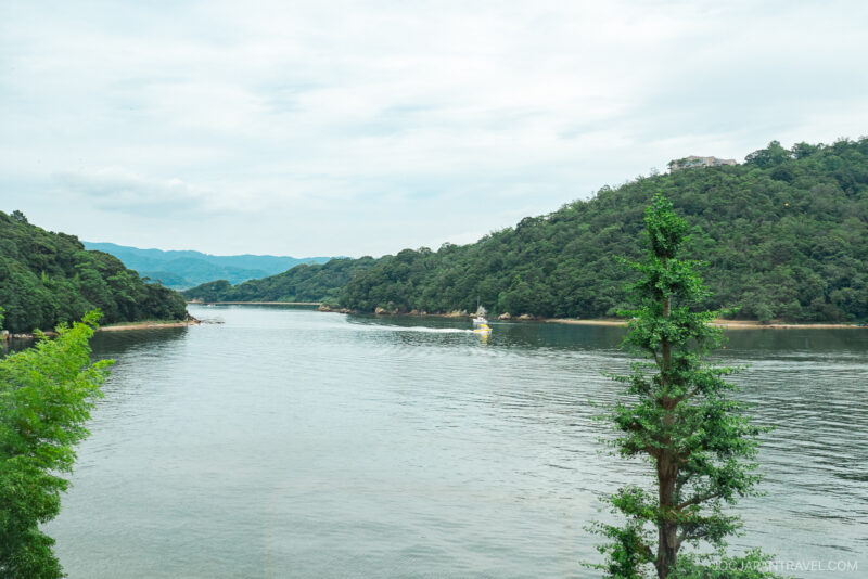 A calm Lake Hamana is surrounded by green, tree-covered hills under a cloudy sky, with a small white boat on the water and a tall tree in the foreground.