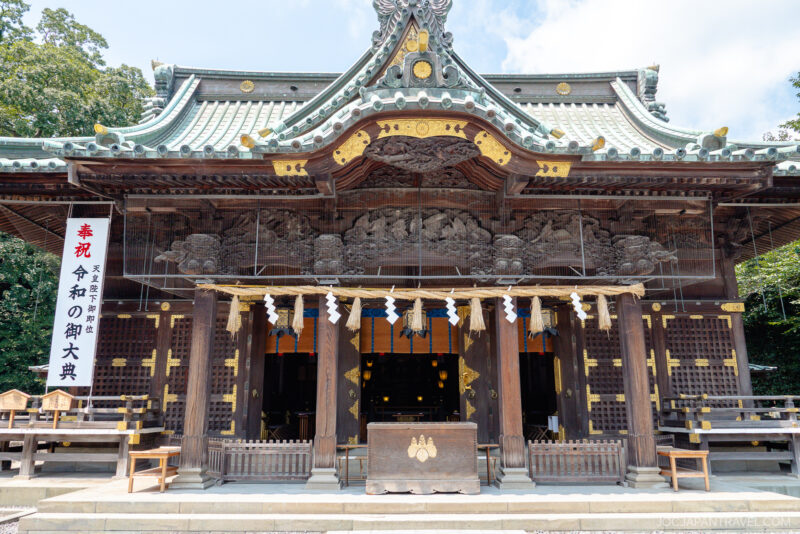 A traditional Japanese Shinto shrine, Mishima Taisha Shrine, features ornate wooden carvings and a green-tiled roof under a blue sky. A banner with Japanese text hangs on the left side of the building.
