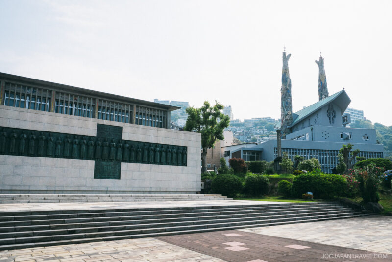 A modern memorial with bronze relief sculptures stands near the Museum of the 26 Martyrs, alongside a building with tall, decorated columns, surrounded by green landscaping and city buildings beneath a bright sky.