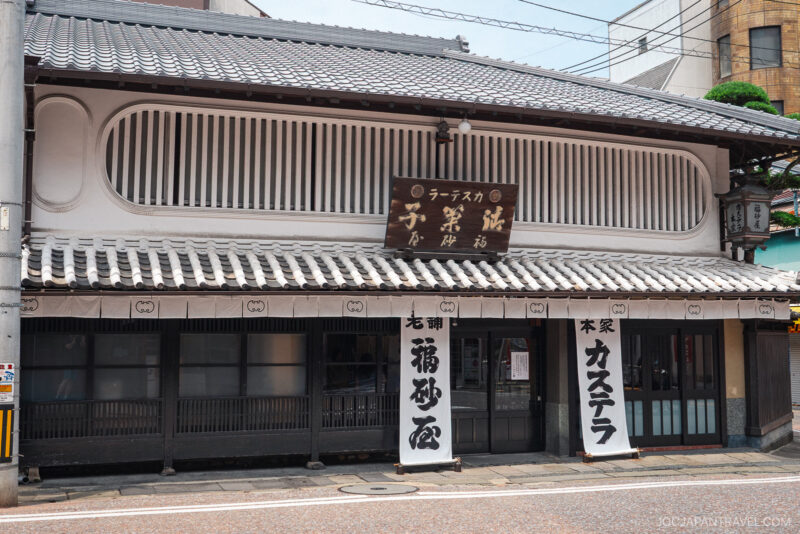 Traditional Japanese storefront with a tiled roof, wooden lattice windows, and white banners with Japanese writing. A large sign above the entrance advertises Nagasaki Castella Cake, inviting visitors to try this famous sweet.
