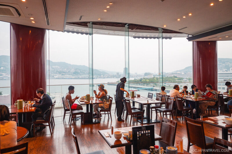A spacious restaurant with large glass windows overlooks a cityscape and waterfront. Diners enjoy Nagasaki noodles at wooden tables, some talking and eating, while a server attends to guests. The atmosphere is bright and relaxed.