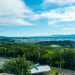 New Furano Prince Hotel (新富良野プリンスホテル) – A Comfortable Stay Near the Flower Fields View of a lush green landscape with tennis courts and buildings in the foreground, dense forests, rolling hills, and an expansive blue sky with scattered clouds—likely taken from the New Furano Prince Hotel’s elevated vantage point.