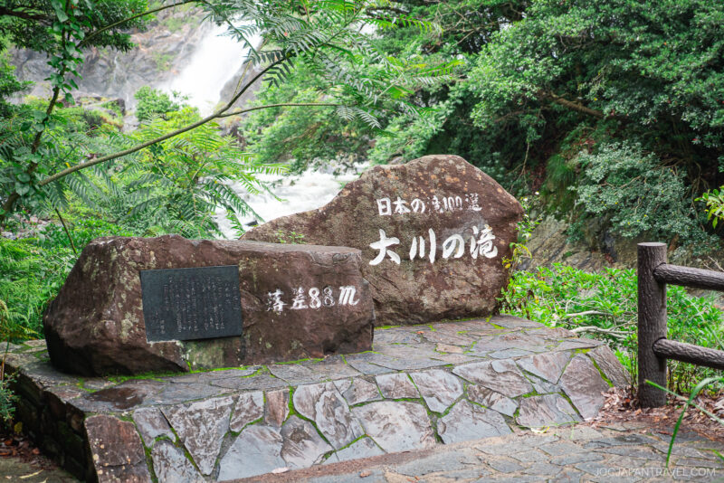 Large stone monument with Japanese inscriptions stands on a stone platform surrounded by lush greenery and trees, with Ohko Waterfall visible in the background. A wooden railing is on the right side.