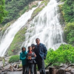 Ohko Waterfall (大川の滝) – Yakushima’s Majestic 88-Meter Cascade Three people in rain jackets stand together in front of Ohko Waterfall, its powerful cascades surrounded by lush green trees and rocky terrain. Mist from the waterfall fills the background.