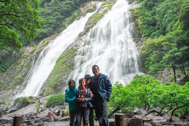 Three people in rain jackets stand together in front of Ohko Waterfall, its powerful cascades surrounded by lush green trees and rocky terrain. Mist from the waterfall fills the background.
