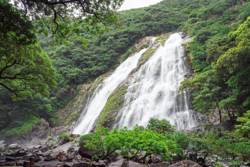 The large, powerful Ohko Waterfall cascades down a green, forested mountain, surrounded by lush trees and rocks under a cloudy sky.