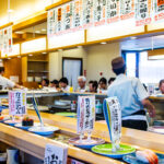 Sapporo Conveyor Belt Sushi – Fresh and Fun Family Dining A busy Conveyor Belt Sushi restaurant with customers seated at the counter. Plates of sushi glide by, while chefs work behind the counter. Japanese menu signs hang above, displaying dish names and prices.