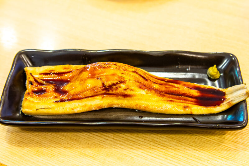 A rectangular black plate, like those at Conveyor Belt Sushi spots, holds a glazed grilled eel fillet topped with a shiny brown sauce and a small dollop of wasabi, set on a light wooden table.