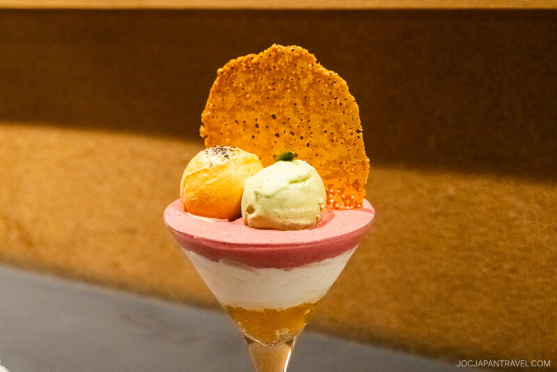 A parfait glass filled with layered mousse and topped with two ice cream scoops, a thin crispy tuile, and herbs—this elegant treat could grace the pages of any Sapporo Sweets Guide, set against a neutral brown background.