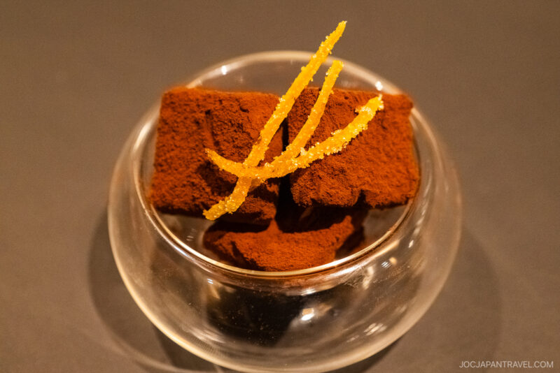 Glass bowl of chocolate truffles dusted with cocoa powder, topped with candied orange peel strips, featured in the Sapporo Sweets Guide, set on a dark surface.