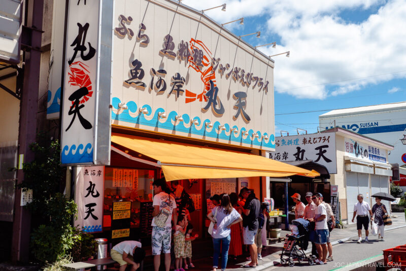 People stand in line outside a Numazu seafood restaurant with yellow awnings and large signs featuring Japanese text and a red fish graphic, all under a bright blue sky.