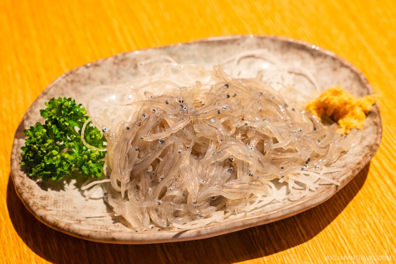 A plate of fresh raw shirauo (whitebait fish) from Numazu served on shredded daikon radish, garnished with parsley and a small mound of ginger paste, arranged on a ceramic dish atop a wooden table.